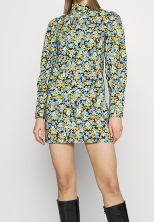Femme portant une mini-robe à manches longues et col haut avec un motif floral dense bleu, jaune et blanc, associée à des bottes noires montantes jusqu'aux genoux.