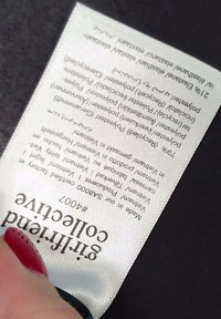 Une main tient une étiquette de vêtement blanche avec la composition détaillée du tissu et des informations sur le produit sur un fond noir.