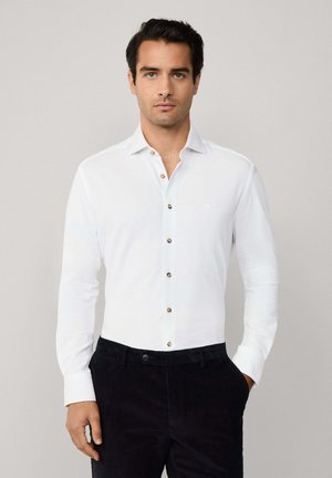 Camisa blanca de manga larga con botones marrones, fabricada en un tejido suave y con un cuello clásico, combinada con pantalones oscuros. Branding mínimo visible.