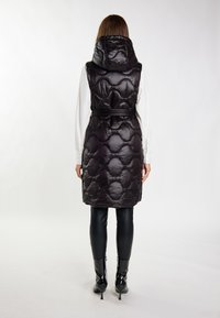 Gilet imbottito nero con design trapuntato, colletto alto e vita cintata, indossato sopra una camicia bianca e abbinato a leggings neri e stivaletti.