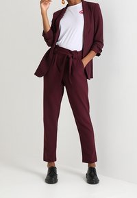 Bordeaux blazer met bijpassende broek; getailleerde pasvorm, gladde stof. Witte t-shirt eronder met een klein rood logo. Zwarte schoenen.