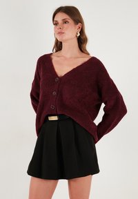Burgundi színű, oversized pulóver V-nyakkal, három gombbal és puha textúrával, fekete, pliszírozott szoknyával és vékony övvel párosítva.