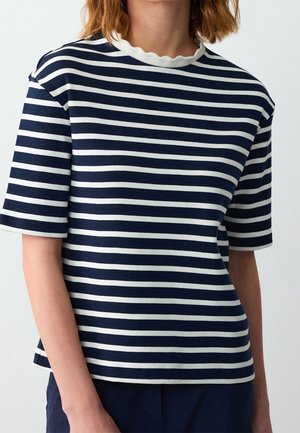 Person, die ein marineblau-weiß gestreiftes Kurzarmshirt mit einem geschwungenen Rundhalsausschnitt vor neutralem Hintergrund trägt.