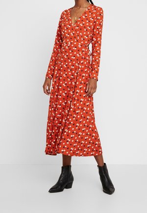 Nainen, joka pukeutunut pitkähihaiseen oranssiin kietaisumekkoon, jossa on pieniä valkoisia ja mustia kukkakuoseja, yhdistettynä mustiin nilkkureihin ja kerroksellisiin kultaiseen kaulakoruihin.