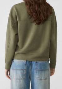 Olijfgroene sweatshirt met een relaxed fit, ronde halslijn en geribbelde manchetten, gecombineerd met lichtblauwe spijkerbroek met twee achterzakken.