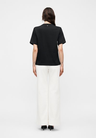 Γυναίκα με την πλάτη, φοράει μαύρο κοντομάνικο μπλουζάκι με το γράμμα "VB" στον λαιμό και φαρδιά λευκά παντελόνια, στέκεται μπροστά σε απλό φόντο.