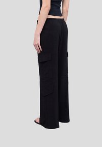 Zwarte cargo pants met wijde pijpen, twee zijzakken, een elastische tailleband en een chevronsteekdetail aan de zijkanten. Zacht textuurmateriaal.