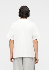 Άτομο με σκούρα μαλλιά μέχρι τους ώμους που φοράει απλό λευκό υπερμέγεθες μπλουζάκι και ανοιχτό γκρι παντελόνι, κοιτάζοντας προς τα πίσω σε λευκό φόντο.