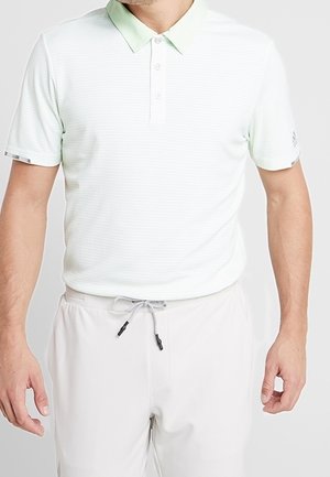 Man draagt een wit polohemd met korte mouwen en een lichtgroene kraag en witte broek met trekkoord, staand met ontspannen armen.
