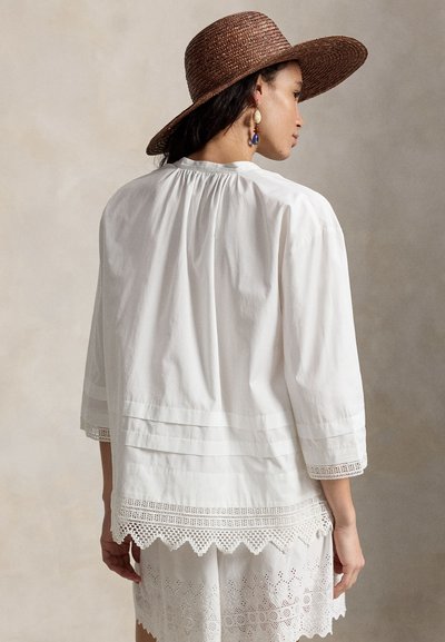 Γυναίκα που φοράει καπέλο καφέ με πλατύ γείσο, άσπρη φαρδιά μπλούζα με δαντελένιο τελείωμα και άσπρες σορτς με κέντημα με οφθαλμότρυπες, κοιτάζοντας πλευρικά μπροστά σε ουδέτερο φόντο.