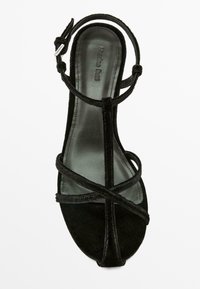 Sandale en daim noir avec un bout ouvert, des brides croisées, une boucle à la cheville et une doublure intérieure lisse. Talon plat avec une forme de bout arrondie.