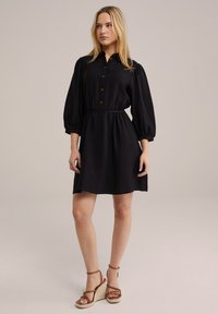 Rochie neagră cu guler, mâneci bufante; material texturat, talie elastică și tiv până la genunchi. Asortată cu sandale cu brâu de culoare bej.