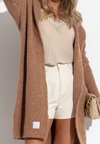 Langer, gestrickter Cardigan in Hellbraun mit Rippstruktur, getragen über einem beigen Oberteil und weißen Shorts. Besitzt ein Etikett am Ärmel.