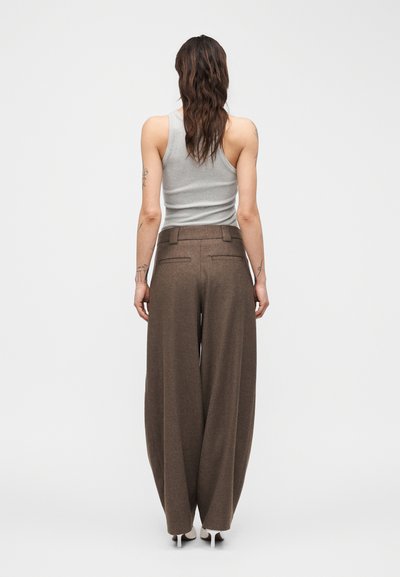 Γυναίκα με μακριά κυματιστά μαλλιά, φορά γκρι αμάνικο τοπ, φαρδιά καφέ παντελόνια και λευκές γόβες, στέκεται με την πλάτη προς το μέρος μας μπροστά σε ένα απλό φόντο.