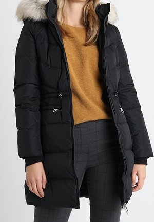 Kobieta ubrana w czarną pikowaną zimową kurtkę z kapturem obszytym futrem, musztardowy sweter i szare spodnie w kratę, stojąca na białym tle.