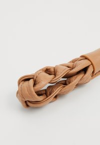 Accessoire en cuir tressé de couleur marron clair, présentant une texture lisse et une courbure homogène, fixé par un bandeau assorti.
