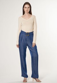Beige Langarmshirt mit V-Ausschnitt, kombiniert mit lockeren blauen Hosen, die einen breiten blauen Gürtel mit goldfarbenen Beschlägen und Ösen-Details aufweisen.