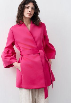 Γυναίκα με σγουρά σκούρα μαλλιά που φοράει ένα φωτεινό ροζ σακάκι με ζώνη, φαρδιά μανίκια και υφασμάτινο, ανοιχτόχρωμο παντελόνι με υφή.