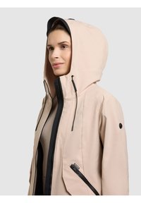 Ženska, oblečena v bež kapucar s črnim zadrgo in vrvicami, obračena levo na enostavni podlagi.