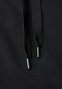 Dettaglio di una felpa nera con cappuccio che presenta una morbida texture del tessuto, spesse corde nere e aglette metalliche alle estremità.