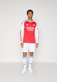 Rød og hvid langærmet fodboldtrøje med et struktureret mønster, der har "Emirates" logoet, parret med hvide shorts og sokker med blå striber.