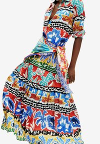 Femme portant une robe fleurie aux motifs éclatants, manches retroussées, avec un foulard coloré noué ceinturant la taille.