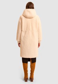 Kremowy, teksturowany płaszcz z polaru z kapturem, z długimi rękawami i luźnym krojem, sięgający do połowy łydki.