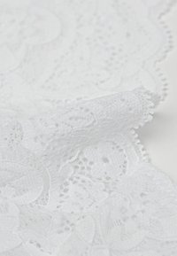 Witte kanten stof met een bloemenpatroon en uitgesneden details, afgewerkt met golvende randen, die complexe texturen en een zachte afwerking toont.