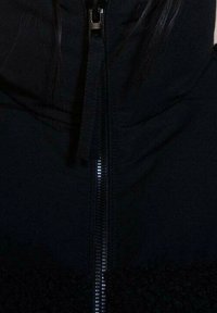 Zbliżenie na czarną kurtkę zapinaną na suwak z widocznym suwakiem oraz fragmentem długich ciemnych włosów po bokach.