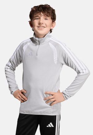 Teenager-Junge mit lockigem Haar lächelt, trägt ein hellgraues Adidas Langarmshirt und schwarze Adidas-Hose, die Hände in die Hüften gestützt vor schlichtem Hintergrund.