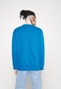 Sudadera azul brillante con cuello redondo y mangas largas, combinada con vaqueros azules claros. Presenta una textura suave y un ajuste relajado.