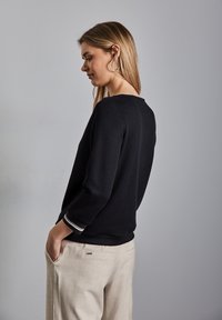 Schwarzer, strukturierten Pullover mit Dreiviertelärmeln, der mit gestreiften Bündchen versehen ist, kombiniert mit hellbeigen Hosen. Sauberes und einfaches Design.