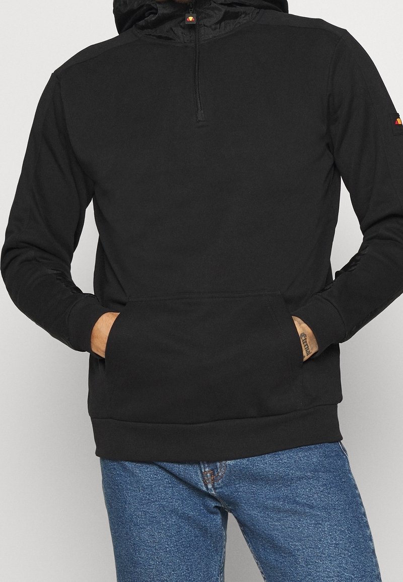 Sweat à capuche noir avec une poche avant et une courte fermeture éclair, porté avec un jean bleu, les mains rangées dans la poche.