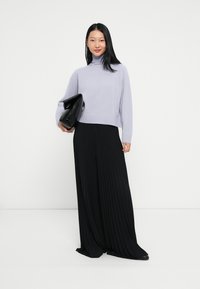 Hosszú fekete hajú nő levendula színű garbót és fekete pliszírozott bő szárú nadrágot visel, fekete clutch táskát tart a kezében.