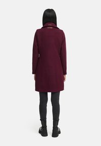 Bordeaux wollen jas met een gestructureerde silhouet, reverskraag en twee accentdetails op de achterkant, gecombineerd met zwarte skinny jeans en laarzen.