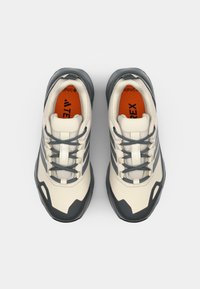 Zapatillas deportivas en beige y gris oscuro con parte superior texturizada, cierre de cordones, plantilla naranja y detalles de logotipo distintivos. Suela de goma redondeada.
