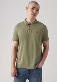 Polo din bumbac de culoare verde măsliniu, cu guler, trei nasturi și un mic logo roșu Levi's pe piept. Purtați cu blugi din denim bleu deschis.