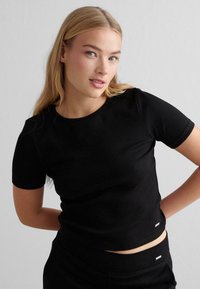Schwarzes, tailliertes Kurzarmshirt mit geripptem Muster und rundem Halsausschnitt. Verfügt über ein kleines Logo am Saum. Kombiniert mit passenden Bottoms.