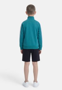 Αγόρι που φοράει τιρκουάζ μπουφάν με λευκές ρίγες στα μανίκια, μαύρο σορτς και λευκά αθλητικά, στέκεται πλάτη σε λευκό φόντο.