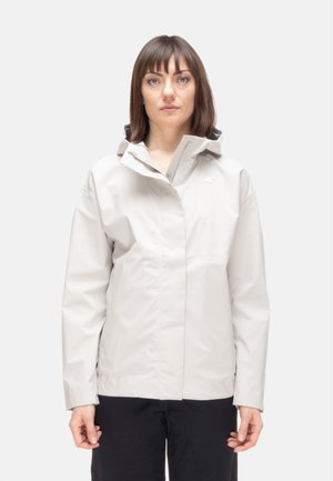 Donna con capelli castani lisci che indossa una giacca antipioggia grigia chiaro con cappuccio e pantaloni neri, rivolta in avanti su uno sfondo bianco semplice.