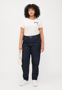 Curly-haired person stands front-facing, wearing a white tee, dark jeans, and white sneakers, holding a small beige handbag.