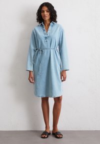 Robe en denim bleu clair avec un col, des manches longues et une ceinture à nouer à la taille. Présente un ourlet arrondi et des coutures visibles. Portée avec des sandales noires à enfiler.