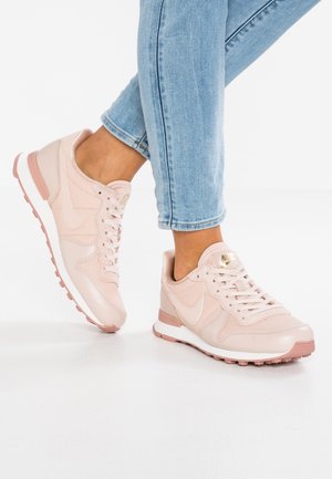 Osoba ubrana w jasnoniebieskie dżinsy i pastelowe różowe sneakersy Nike z białymi podeszwami, stojąca na białej podłodze na jednolitym tle.