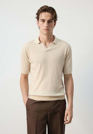 Joven con piel clara y cabello castaño que lleva una camiseta polo beige acanalada de manga corta y pantalones marrón oscuro, de pie frente a un fondo liso.