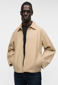 Μπεζ, ελαφρύ μπουφάν με φερμουάρ μπροστά, μυτερό γιακά και πλαϊνές τσέπες, φορεμένο πάνω από μαύρο πουκάμισο, σε ουδέτερο φόντο.