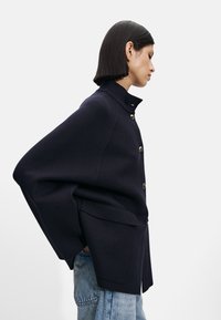 Mørkeblå ullfrakk med høy krage og gullknapper, med vide ermer og en strukturert fasong, kombinert med lyseblå denimjeans.