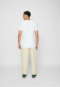 Weißes Baumwoll-T-Shirt mit Rundhalsausschnitt, kombiniert mit hellbeigen, locker sitzenden Hosen und grünen Schuhen, von hinten betrachtet.