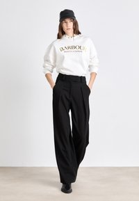 White sweatshirt with "BARBOUR INTERNATIONAL" in gold lettering, paired with black wide-leg trousers and a black cap. Smooth textures, relaxed fit.