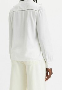 Vit långärmad blus med knappad krage, subtila veck vid axlarna och dekorativ sömnad över den övre ryggen. Bärs med ljusa byxor.