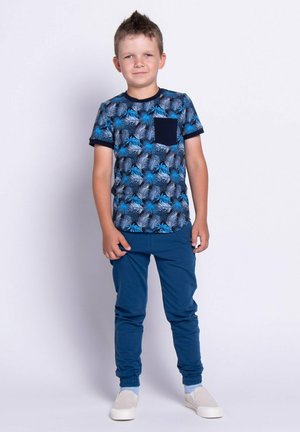 Niebieska koszulka z wzorem, z krótkim rękawem i kieszenią na piersi, zestawiona z ciemnoniebieskimi zwężanymi spodniami oraz jasnymi butami wsuwanymi.